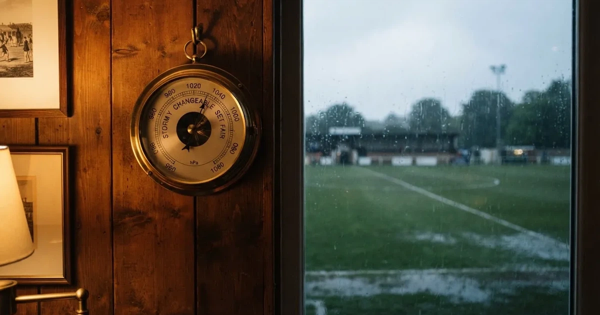 Ein Wetterbarometer hängt an einer Holzwand neben einem Fenster, draussen liegt ein nasser Fussballplatz im Regen.