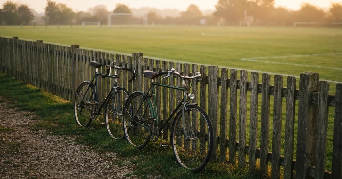 Drei Fahrräder lehnen nebeneinander an einem Zaun vor einem Sportplatz, dahinter ein grüner Rasen im Dunst.
