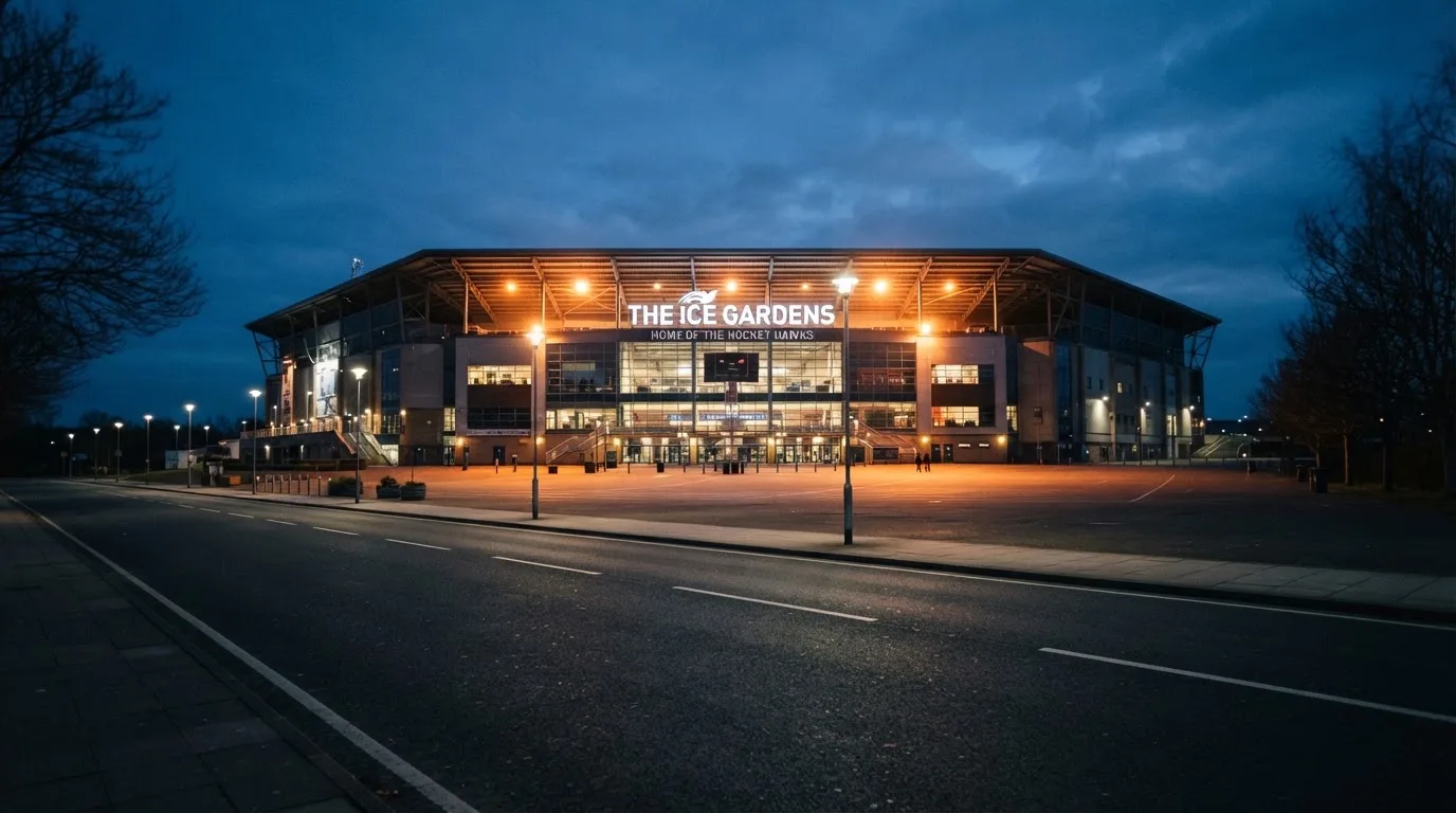Ein grosses Eisstadion aus der Ferne gesehen, hell erleuchtet von Flutlicht in der Abenddämmerung, davor eine leere Strasse.