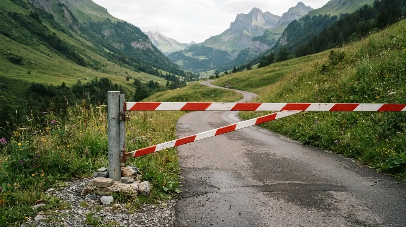 Ein Schlagbaum sperrt eine Landstrasse in den Bergen, dahinter windet sich der Weg zwischen grünen Hängen.