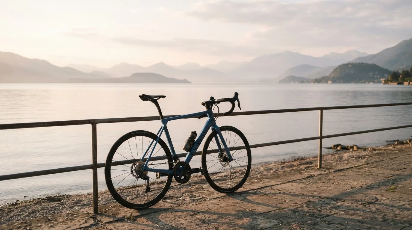 Ein Rennrad lehnt an einem Geländer am Seeufer, dahinter liegt ruhiges Wasser vor einer Bergkette.