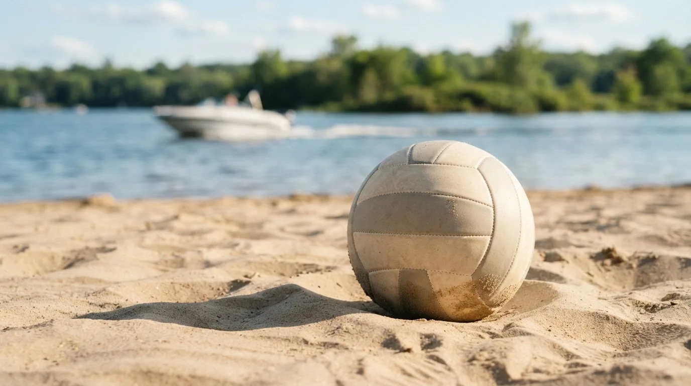 Ein Volleyball liegt im Sand eines Spielfelds am Seeufer, im Hintergrund gleitet ein Boot über das Wasser.