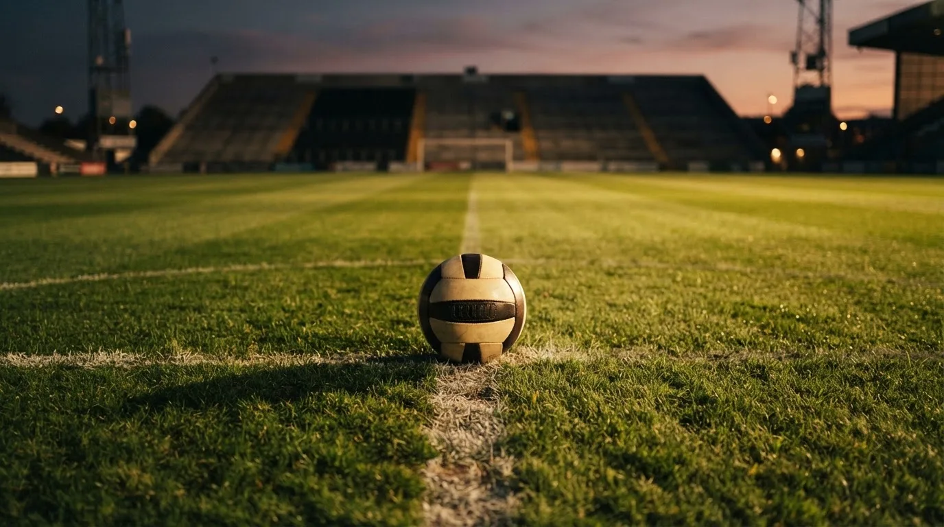 Ein Fussball liegt auf dem Mittelkreis eines leeren Rasenplatzes, dahinter ragen leere Tribünenreihen in den Abendhimmel.
