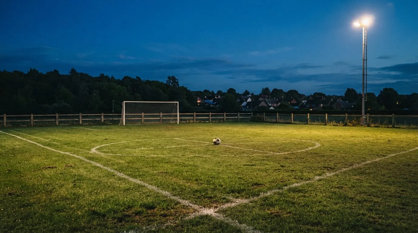 Ein kleiner Dorf-Fussballplatz liegt in der Dämmerung unter einem hohen Flutlichtmast, am Spielfeldrand steht ein Holzzaun.