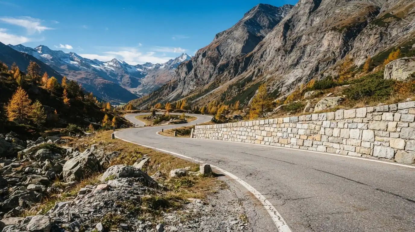 Eine Passstrasse im Hochgebirge säumt eine niedrige Steinmauer, dahinter ragen Felswände auf und der Herbsthimmel ist klar.