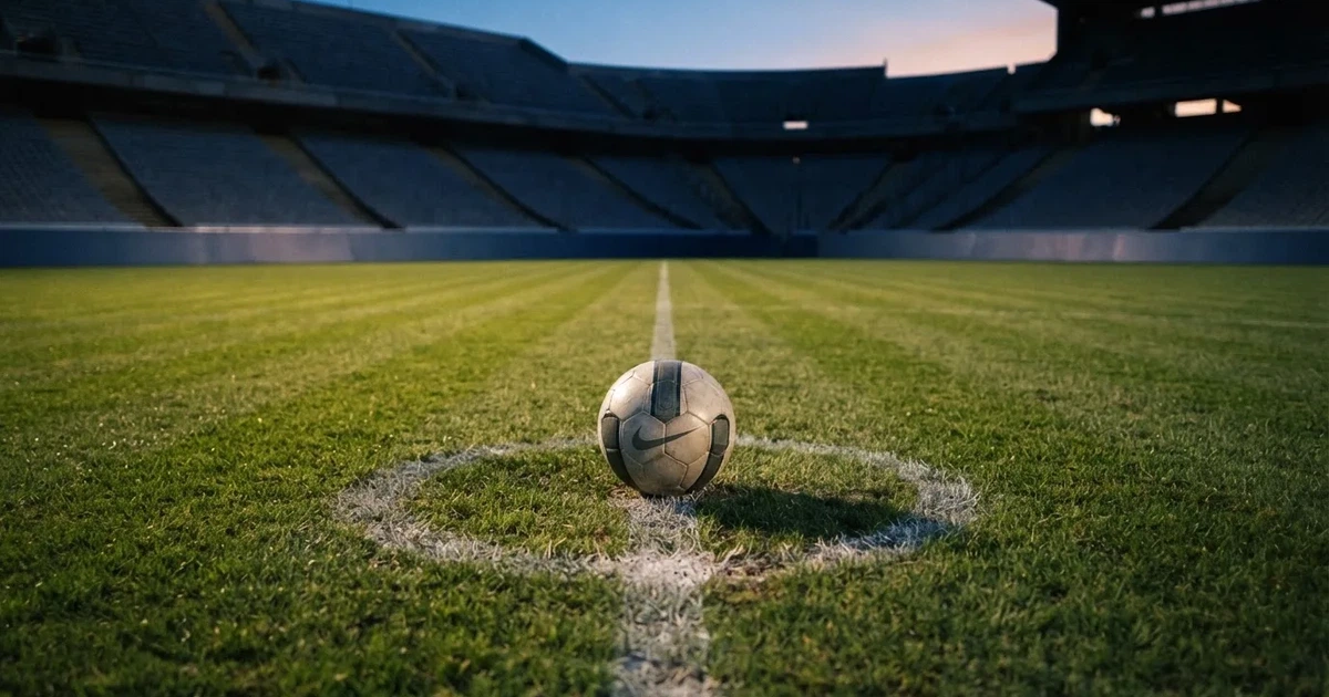 Ein Fussball liegt auf dem Mittelpunkt eines leeren Rasens, im Hintergrund steigen steile Tribünen in der Dämmerung auf.