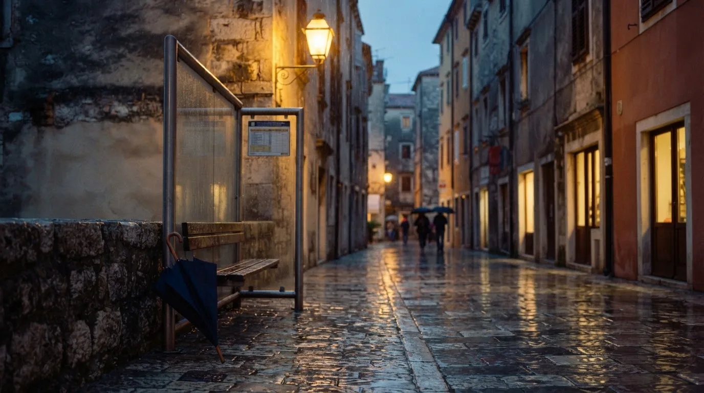 Eine Tramhaltestelle bei Regen in einer Altstadtgasse, das Kopfsteinpflaster glänzt und ein einzelner Regenschirm lehnt an einem Mäuerchen.