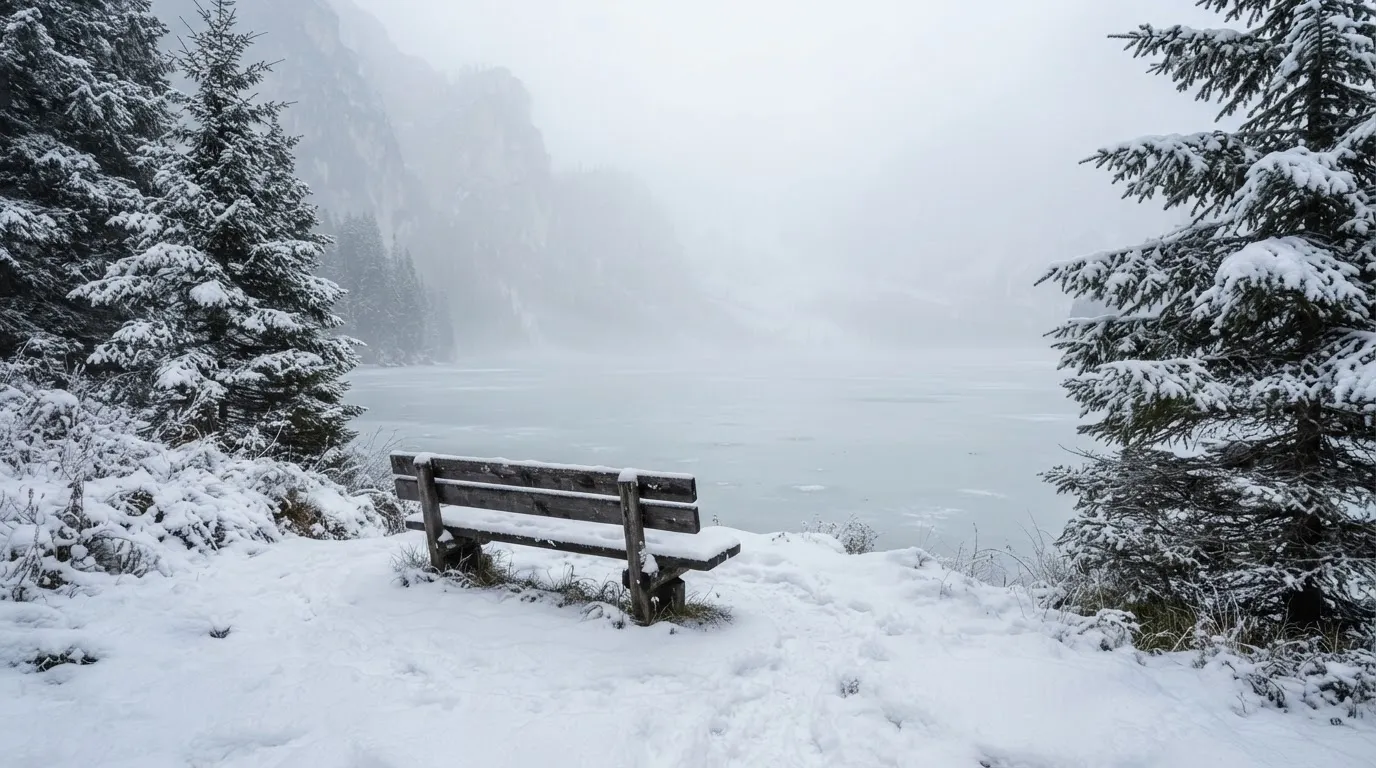 Ein zugefrorener Bergsee im Morgendunst, am Ufer steht eine einsame Holzbank zwischen verschneiten Tannen.