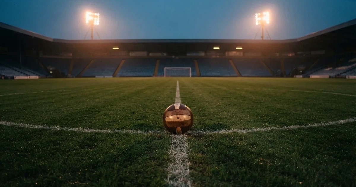 Ein Fussball liegt auf dem Anstosspunkt eines leeren Rasenplatzes in der Abenddämmerung, dahinter leuchten Flutlichtmasten über einer Tribüne.