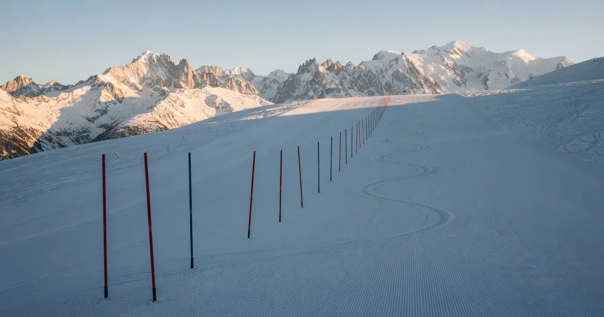 Ein frisch präparierter Hang mit Slalomstangen in einer Reihe im ersten Morgenlicht, Schneespuren im Hang und Berge im Hintergrund.