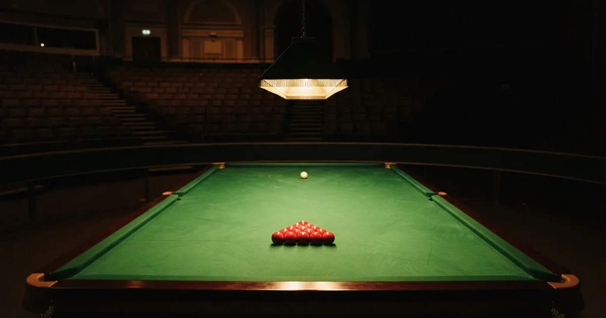Ein Snookertisch mit grünem Tuch und einem Dreieck aus roten Kugeln unter einer tief hängenden Lampe in einer leeren Halle.