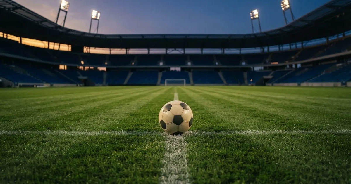 Ein Fussball liegt auf dem Anstosspunkt eines leeren Rasenplatzes, dahinter ragen die Ränge eines grossen Stadions in der Abenddämmerung auf.