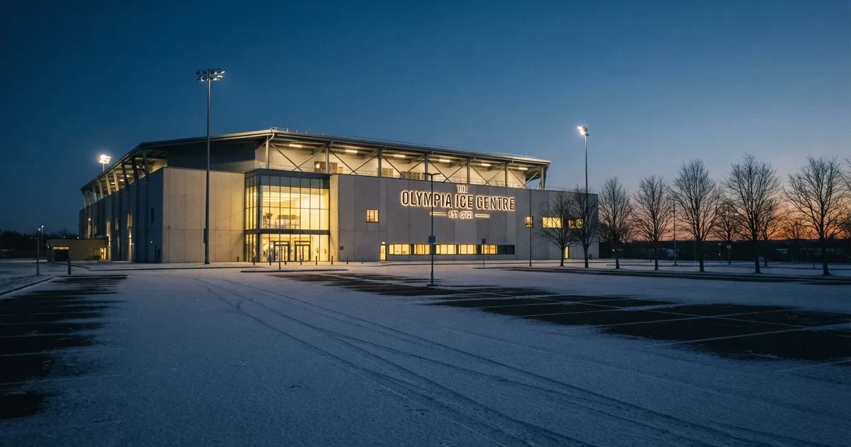 Eine Eishalle steht in der Dämmerung unter Flutlicht, aus der Ferne gesehen, davor ein leerer Parkplatz mit Schnee.