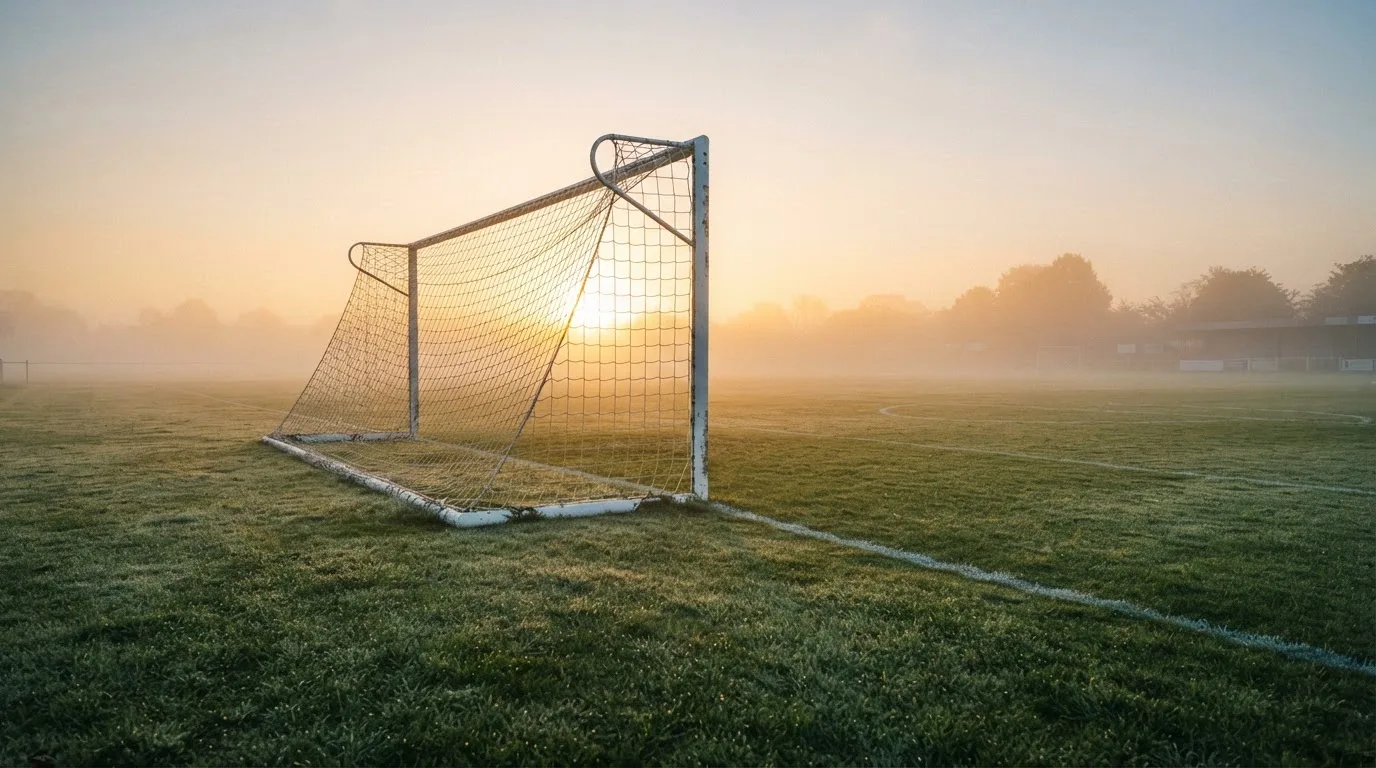 Ein Torpfosten mit Netz steht im Gegenlicht auf einem leeren Fussballplatz, über dem Rasen liegt Nebel.
