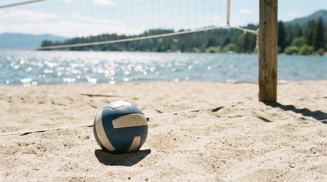 Ein Volleyball liegt auf dem Sand eines Strandfeldes am Seeufer, im Hintergrund glitzert das Wasser.