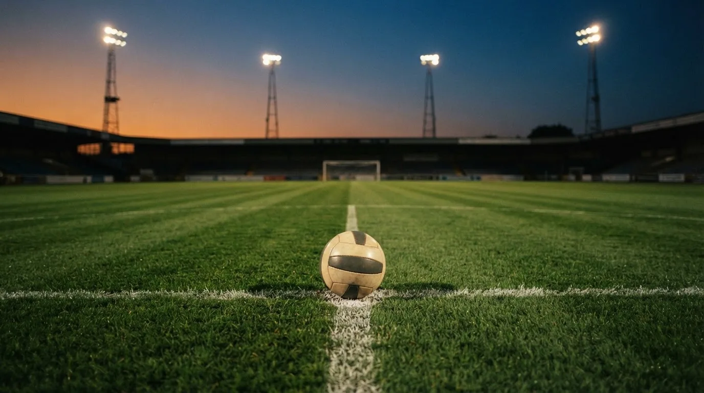 Ein Fussball liegt auf dem Mittelpunkt eines leeren Rasenplatzes, im Hintergrund ragen Flutlichtmasten in die Abenddämmerung