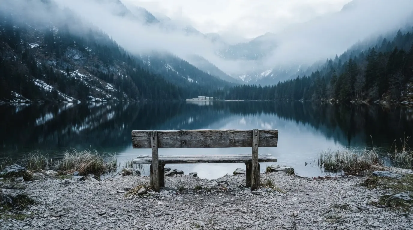 Eine Holzbank am Ufer eines ruhigen Bergsees, am gegenüberliegenden Ufer steigen dunkle Hänge in den Frühnebel
