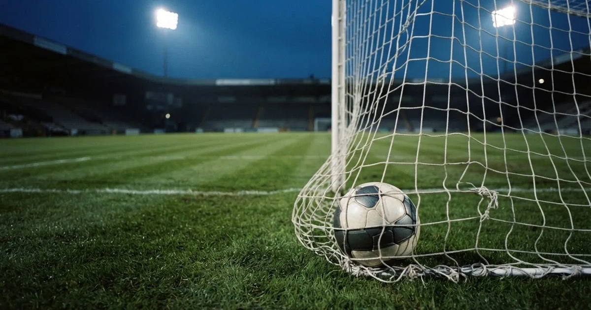 Ein Fussball liegt im Netz eines Tores, Flutlicht und kalte Abendluft liegen über dem leeren Platz