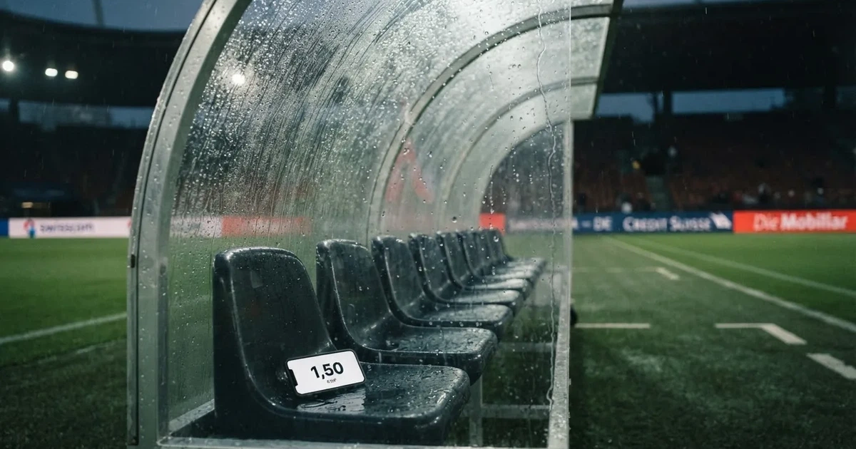 Eine leere Ersatzbank unter einem Plexiglasdach am Spielfeldrand, Regentropfen auf der Scheibe, dahinter ein Rasenplatz