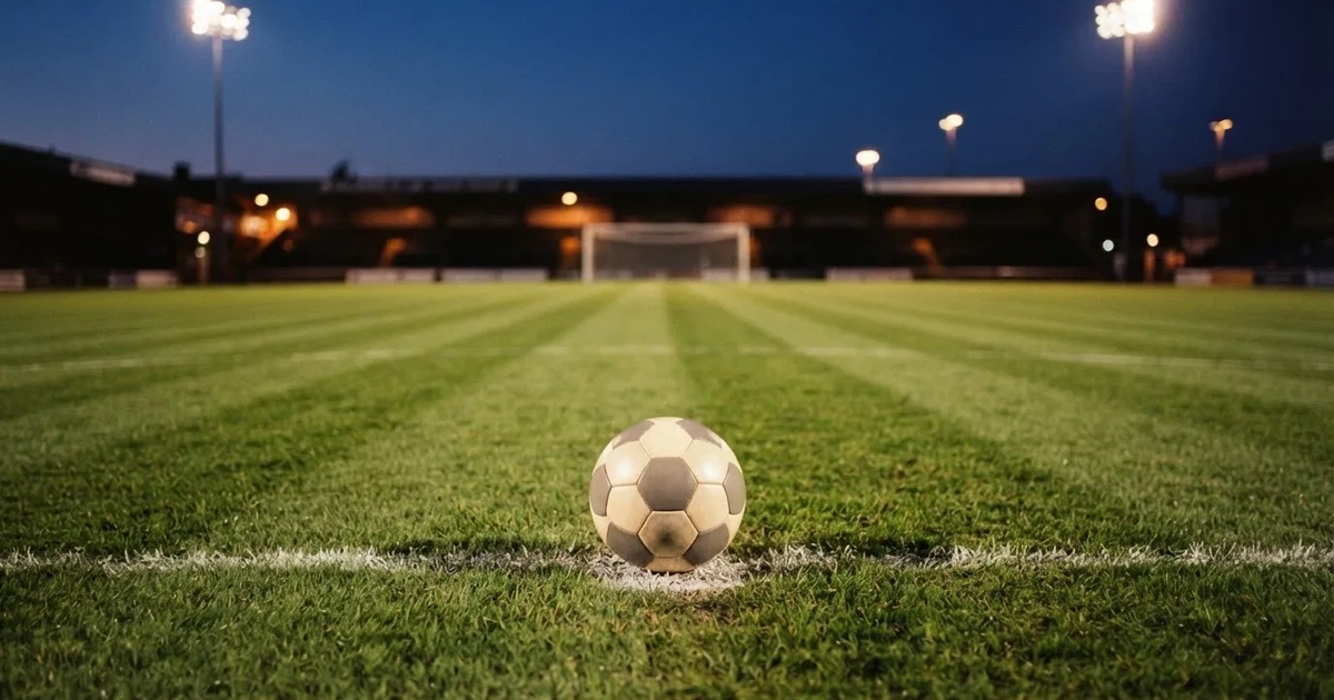 Ein Fussball liegt auf dem Anstosspunkt eines leeren Rasenplatzes in der Abenddämmerung, im Hintergrund leuchten Flutlichtmasten.