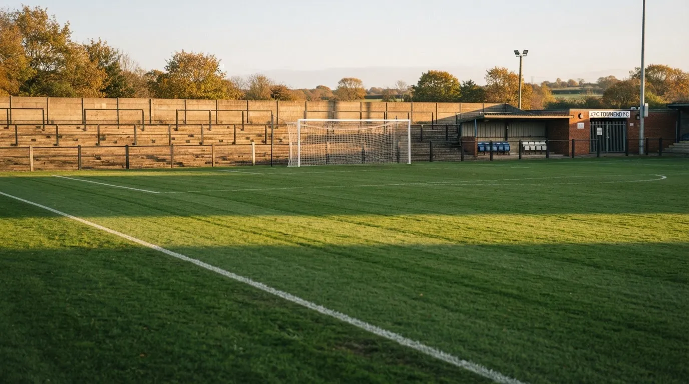 Leere Zuschauerränge eines kleinen Stadions im Herbstlicht, davor ein Rasenplatz mit frisch gezogenen Linien.