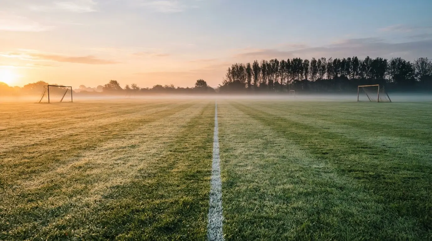 Ein Rasenspielfeld bei Sonnenaufgang, die Mittellinie verläuft quer durchs Bild und Tau glitzert auf dem Gras.