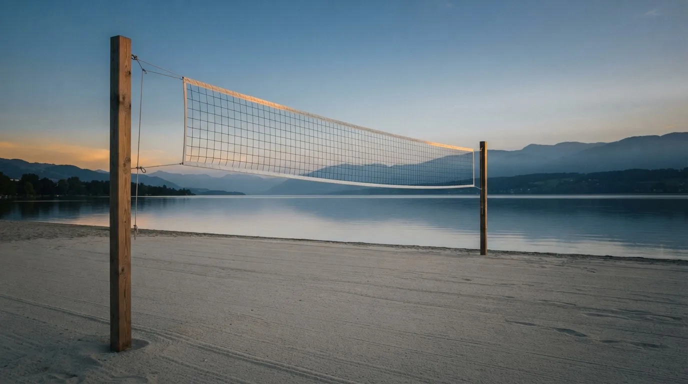 Ein Volleyballnetz steht an einem Seeufer in der Abenddämmerung, der Sand ist leer und das Wasser glatt.