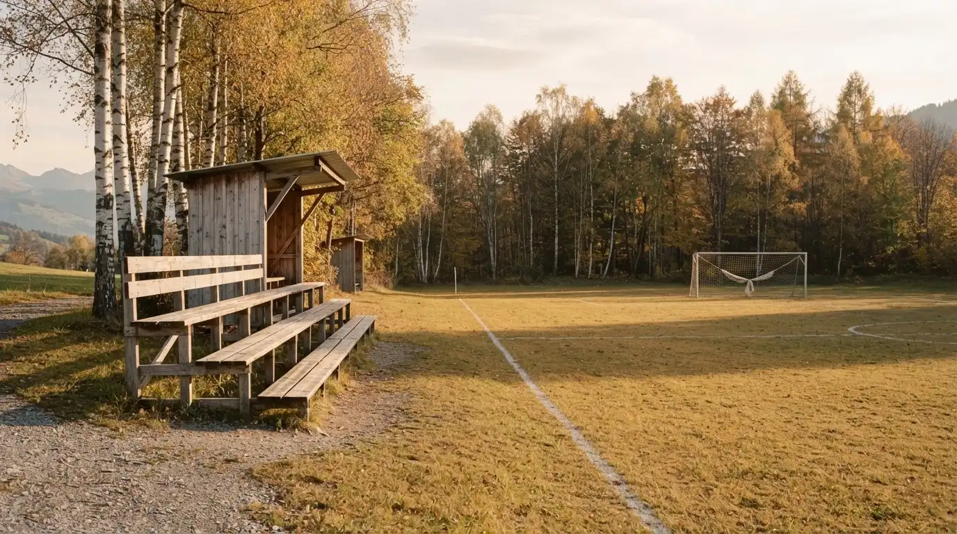 Eine leere Holztribüne steht am Waldrand neben einem kleinen Sportplatz im Herbstlicht.