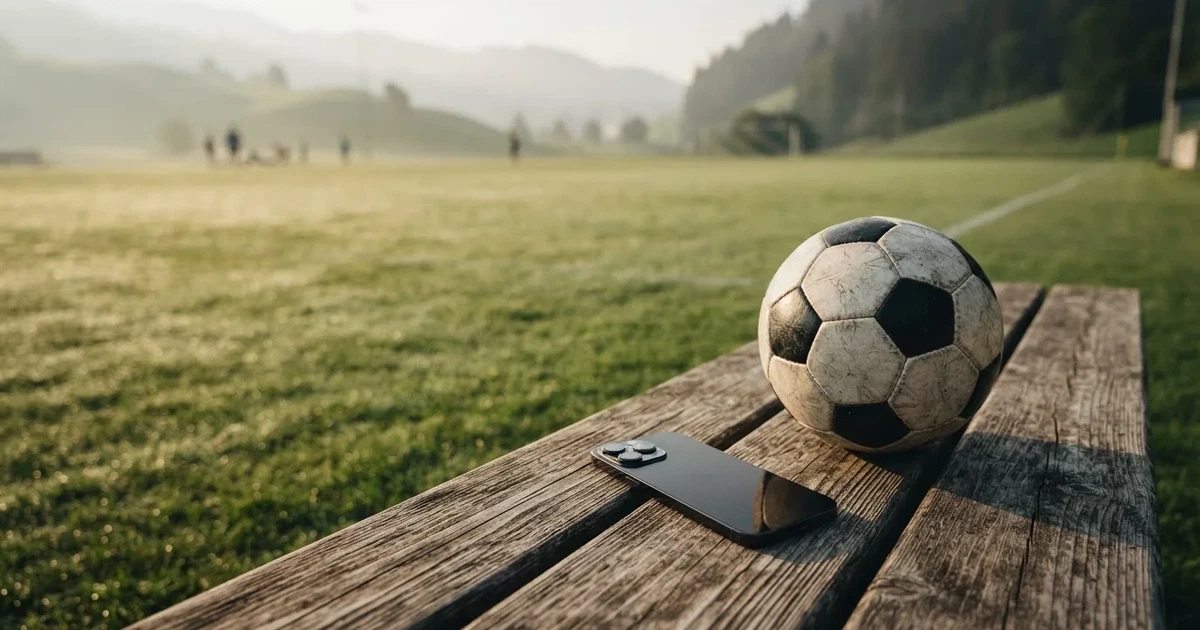 Ein Smartphone mit dunklem Bildschirm liegt neben einem Fussball auf einer Holzbank am Rand eines Trainingsplatzes im Morgenlicht.