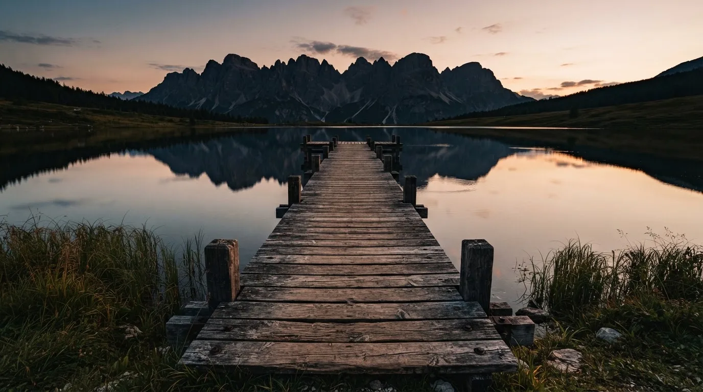 Ein leerer Holzsteg ragt am Abend in einen ruhigen Bergsee, das Wasser spiegelt den Himmel.