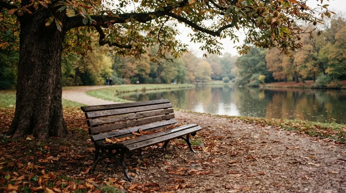 Eine Parkbank unter einer Kastanie, Herbstlaub bedeckt den Kiesweg, im Hintergrund ein stiller Teich.