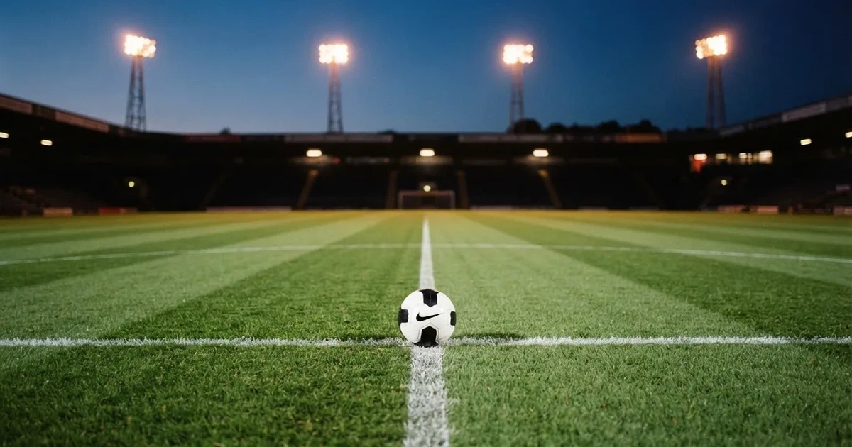 Ein Fussball liegt auf dem Anstosspunkt eines leeren Rasenplatzes, im Hintergrund glimmen Flutlichtmasten im Abendlicht.
