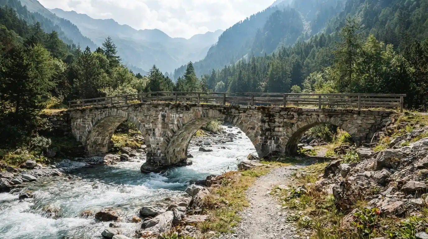 Eine alte Steinbrücke überspannt einen Gebirgsfluss, im Hintergrund steigen bewaldete Hänge an.