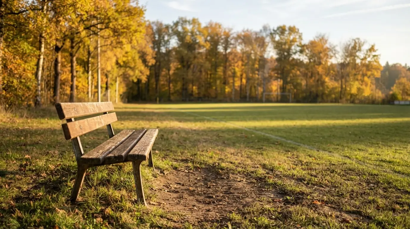 Eine hölzerne Bank steht an der Seitenlinie eines leeren Sportplatzes, dahinter Bäume im Herbstlicht.